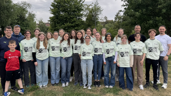 Mannschaftsfoto der Teilnehmenden des Staffellaufs. Zu sehen sind 4 Lehrer der Schule als Läufer und 20 Schülerinnen und Schüler der Jahrgangsstufe 11 in ihren Streckenposten-Shirts