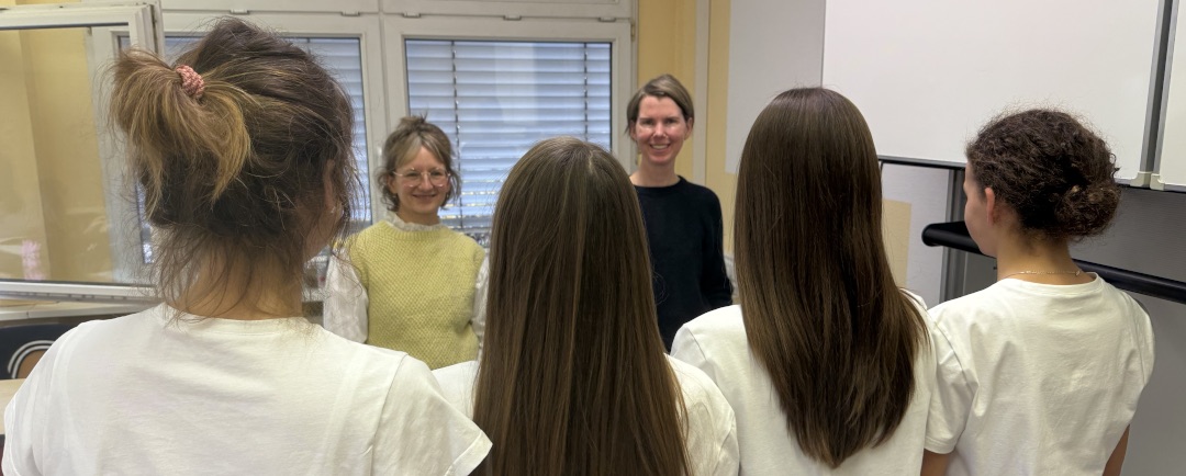 Die beiden Bekleidungstechnikerinnen Anke Beutel und Kathleen Tischer stehen vor Schülerinnen in weißen Shirts.